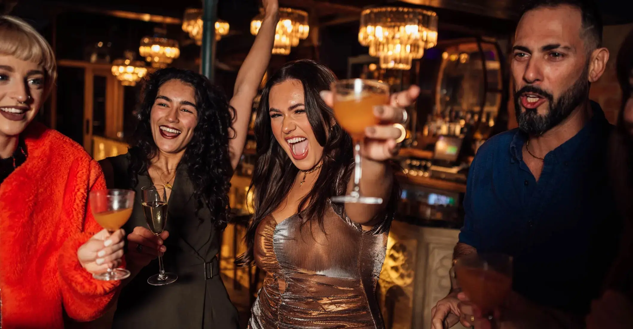 Friends laughing and raising cocktails together at a lively bar